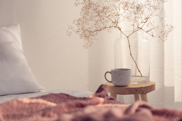 Closeup of coffee cup and flower in glass vase on the bedside table of bright bedroom interior,...