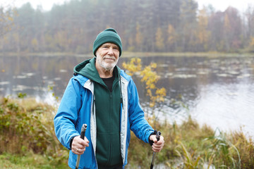 Outdoor shot of happy senior man in warm clothes standing with nordic walking poles in colorful...