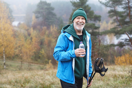 Wellness, health, activity, sports and hobby concept. Picture of happy positive mature bearded male in his seventies posing outdoors in wild nature, holding plastic bottle of water and nordic poles