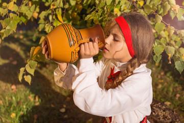 The girl is drinking from a large ceramic jug. The girl is dressed in a national costume.