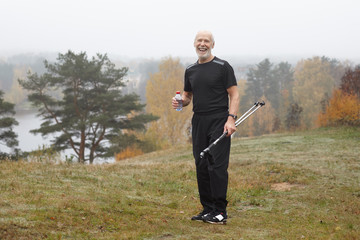 Picture of energetic healthy retired senior man with gray beard posing outdoors with plastic bottle of water, refreshing himself after intensive cardio nordic walking exercise, wearing sports clothes