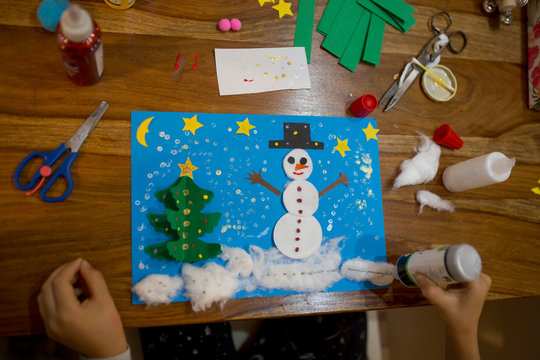Top view of child, doing snowman and winter tree. Scissors, glue stick, pencil, markers, paper sheets and scraps on wooden background.