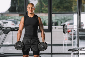 Fototapeta na wymiar smiling african american sportsman looking at camera and exercising with dumbbells at gym