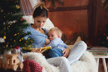 Mother breastfeeding her toddler son sitting in cozy armchair near Christmas tree