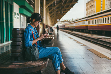 Asian woman traveler used smart phone while wait and check train schedule on the platform of the railway station - travel and transportation concept