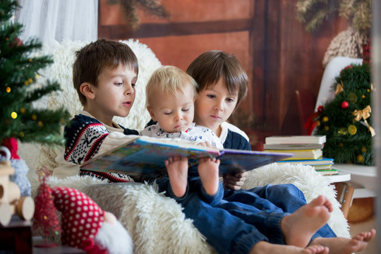 Three children, boy brothers, sitting in rocking chair in cozy living room with christmas decoration, reading a book