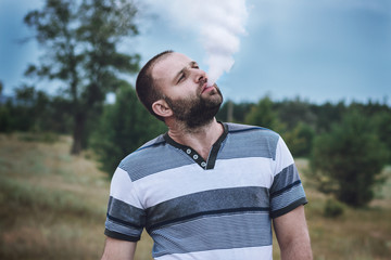 Young man with a beard of Caucasian appearance smokes electronic cigarette in nature, daylight,...