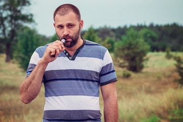 Young man with a beard of Caucasian appearance smokes electronic cigarette in nature, daylight,...