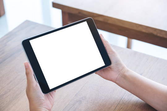 Mockup image of hands holding and using black tablet pc with blank white desktop screen while sitting in office