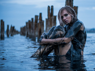 Portrait of Blonde Caucasian Girl Sitting Smeared in a Healthy Black Mud in old Firth with Wooden Posts for Salt Production
