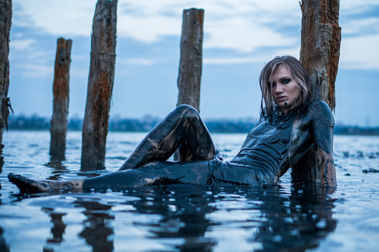Portrait of Blonde Caucasian Girl Lays Smeared in a Healthy Black Mud in old Firth with Wooden Posts for Salt Production