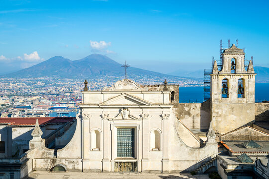 Bright skyline view of Naples, Italy with Mount Vesuvius standing above a cityscape dominated by an old church in the foreground