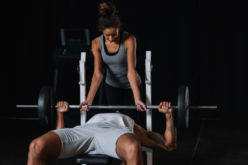 african american woman helping boyfriend to exercising with barbell at gym