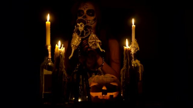 Halloween witch with skull makeup makes voodoo holds knife and wispering spell magic pumpkin chains and candles