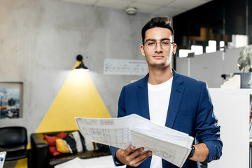 Stylish architect dressed in blue checkered jacket and jeans works with blueprints in the modern office