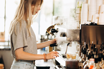 A youthful blonde slim gir,dressed in casual outfit,cleans the coffee machine with steam in a modern coffee shop.
