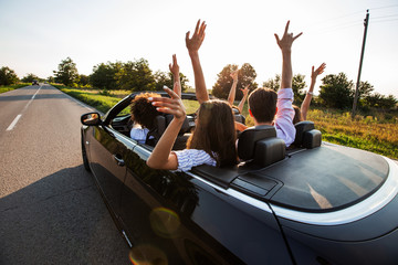 Black cabriolet is on the country road. Happy group of young girls and guys are sitting in the car hold their hands up on a sunny day.