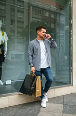 Young man with shopping bags talking on the mobile leaning on a shop window