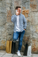 Obraz premium Young man with shopping bags leaning on a stone wall while talking on the mobile