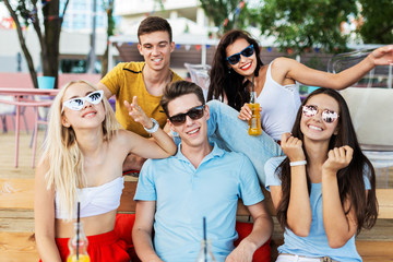 A company of good-looking friends wearing sunglasses laughing and drinking yellow cocktails and socialising at the table in the nice summer cafe. Cheers. Entertainment, having good time. Friendship.