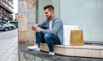 Young man with shopping bags sitting on the curb of a shop window