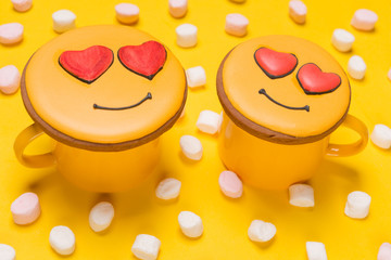 Fototapeta premium minimal concept of love and a date, two yellow cups covered with emoticons-shaped gingerbread with a heart, surrounded by marshmallows, on a yellow background