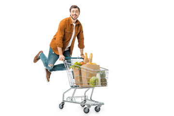 positive man jumping and carrying shopping trolley with products in paper bags isolated on white