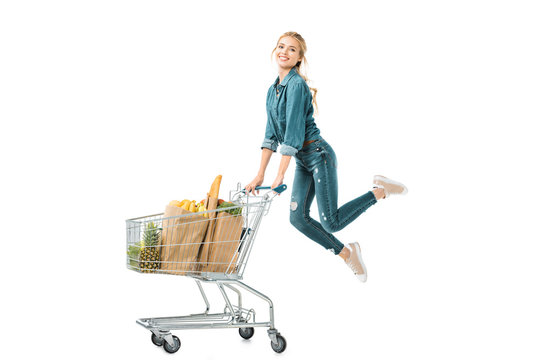 happy young woman jumping near shopping trolley cart with products in paper bags isolated on white