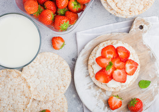 Healthy organic rice cakes with ricotta and fresh strawberries and glass of milk on light stone kitchen background. Top view. Plastic tray of strawberries
