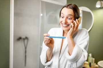 Happy woman in bathrobe excited with a pregnancy test result calling with phone in the bathroom