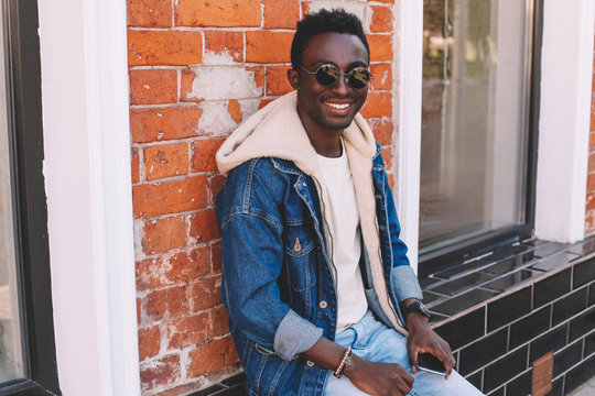 Fashion smiling african man in jeans jacket sits on city street, brick wall background