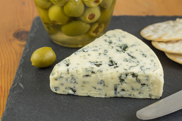 Blue cheese wedge with defocused jar of green olives and crackers in background