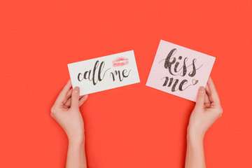 partial top view of person holding cards with kiss me and call me inscriptions and kiss mark isolated on red