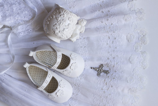 Christening baby shoes, christening cup and crystal cross pendant on vintage lace baptism dress on white background