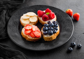 Fresh healthy mini sandwiches with cream cheese, fruits and berries in black plate with cloth. Strawberries, blueberries, bananas and raspberries on stone kitchen table background.Top view.