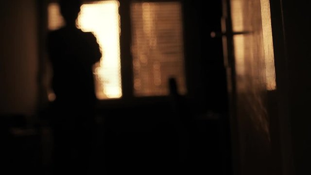 Depressive man in dark room standing by the window, defocussed person in the background