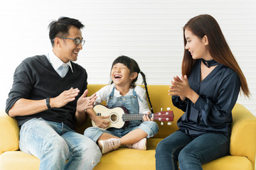 Asian daughter playing guitar and singing with father and mother on sofa in livingroom. concept family enjoyment.