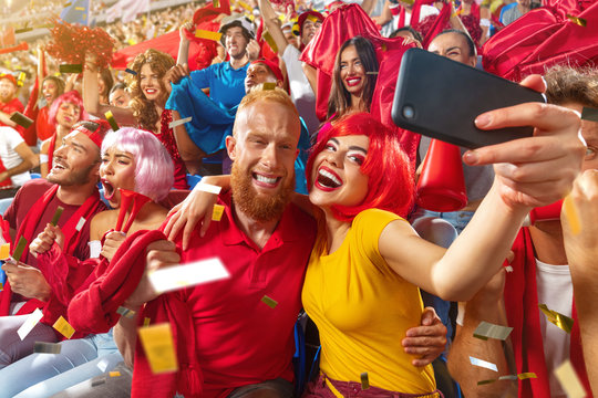 Young sport supporter happy fans at stadium. Beautiful man and woman in a red wig support the football team during the match and making selfie photo