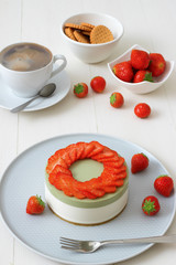 Cooking demo. No-bake two-layered mini strawberry matcha cheesecake. Strawberry slices decorate the top of the cake. 3 cups with coffee, cookies and strawberry in background.