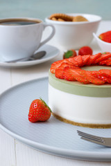 Cooking demo. No-bake two-layered mini strawberry matcha cheesecake. Strawberry slices decorate the top of the cake. 3 cups with coffee, cookies and strawberry in background.