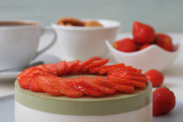 Cooking demo. No-bake two-layered mini strawberry matcha cheesecake. Strawberry slices decorate the top of the cake. 3 cups with coffee, cookies and strawberry in background.