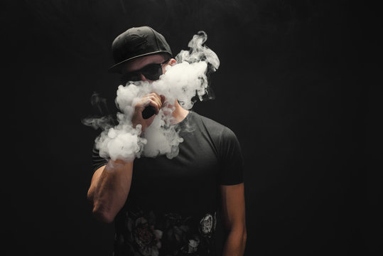 Vape man. Portrait of a handsome young white guy in a modern black cap vaping and letting off puffs of steam from an electronic cigarette 