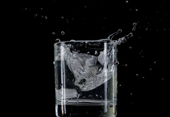 Bounce and splash from a piece of ice thrown into a transparent glass with water on a black background