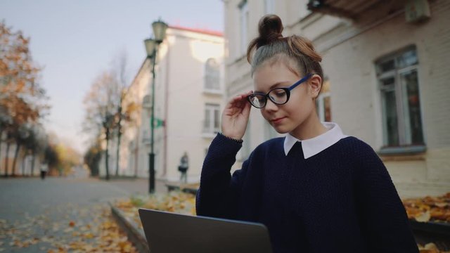 Close up pretty hipster teen girl sitting on a sidewalk on autumn city street and working laptop computer. Schoolgirl using notebook outdoor. Beautiful autumn weather.