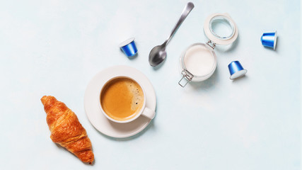 Breakfast concept. Coffee espresso in white cup, coffee beans, capsules, croissants, butter on a blue pastel background with space for text. Flat lay.