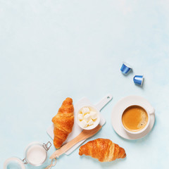 Breakfast concept. Coffee espresso in white cup, coffee beans, capsules, croissants, butter on a blue pastel background with space for text. Flat lay.