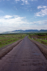 Open dirt road with mountain view, green field and nice cloudy blue sky. Maharashtra, India.