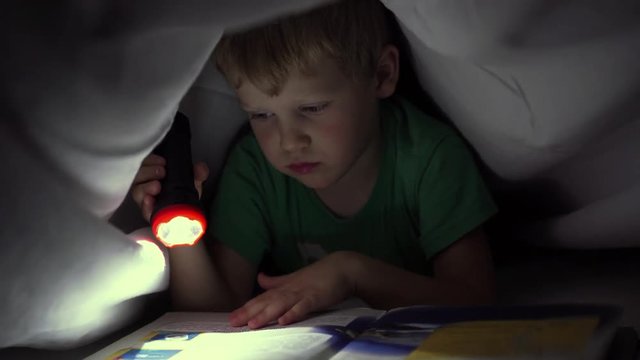 little boy reading a book at night under a blanket with a flashlight