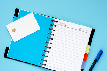 Blank white credit chip card, spiral notebook and a pen on blue background for spending plan concept