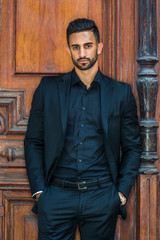 Portrait of Young Handsome East Indian American Businessman with beard in New York, wearing black suit, black shirt, hands in pockets, standing by brown old style office doorway, looking at you..